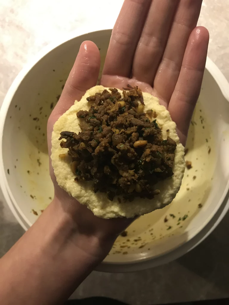 Semolina dough flattened in the palm of a hand and filled with minced meat and pine seeds