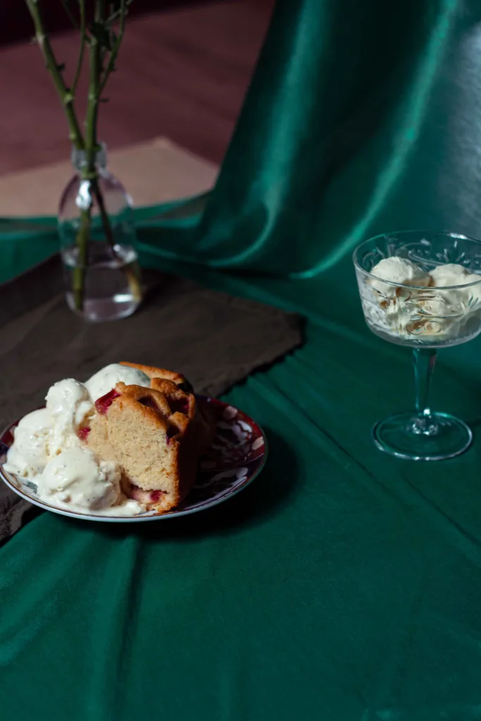 A plate with rhubarb semolina cake and a heap of cardamom ice cream and a champagne coupe glass filled with more ice cream on a green satin cloth.