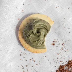 One rooibos shortbread cookie covered in white chocolate matcha ganache, with a bite taken out of it and rooibos tea leaves scattered around it.