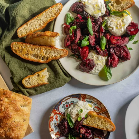 Overhead shot of two plates with balsamic glazed plums and onion, basil, and burrata, and freshly toasted sourdough bread.