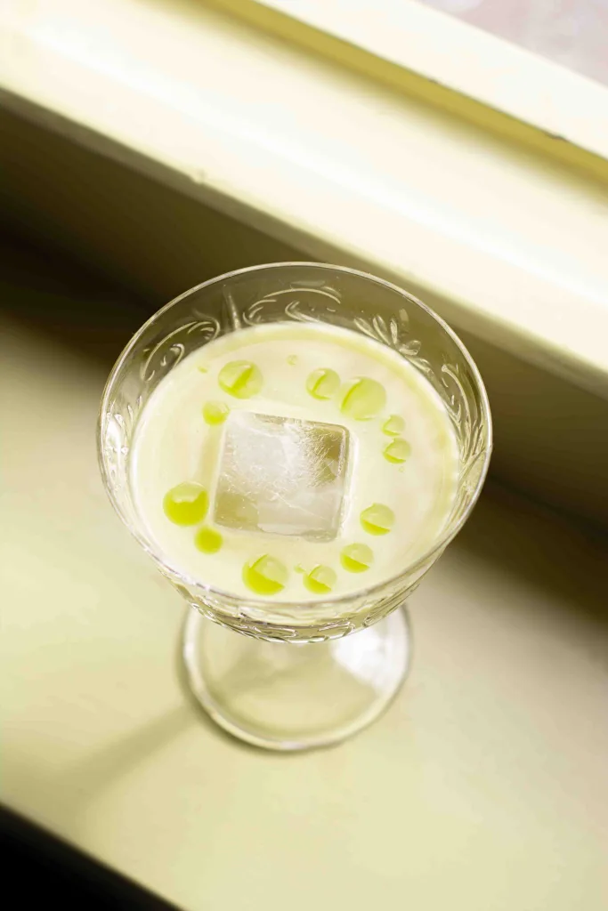 Overhead shot of a champagne coupe glass filled with cream of asparagus consomme, an ice cube, and some drops of tarragon oil standing on a yellow window sill.