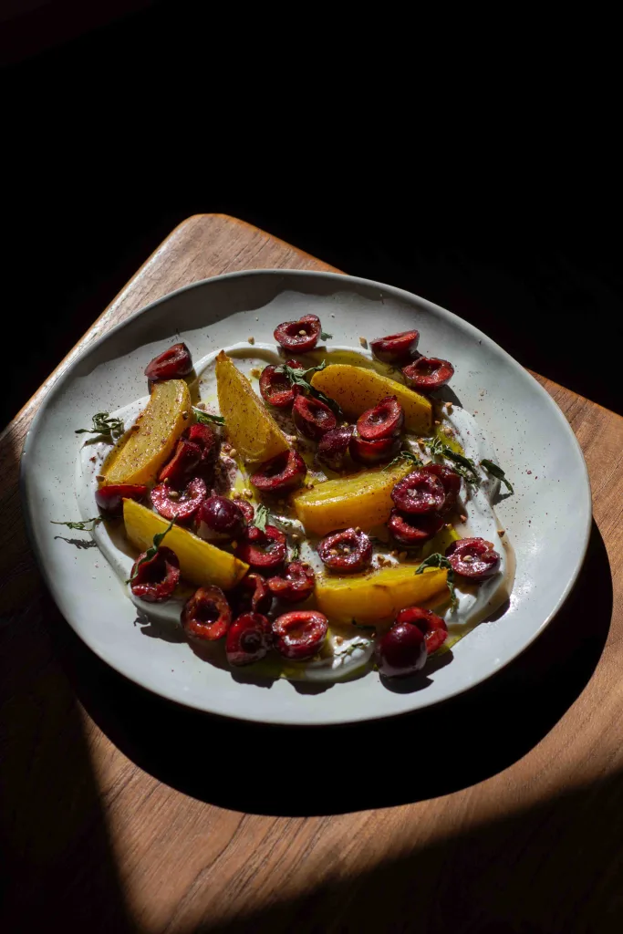 Photo of a plate of whipped goat cheese topped with fresh cherries, roasted beetroot, sumac, and tarragon oil with dark shadows in the background.