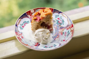 One slice of half-eaten rhubarb lemon rose semolina cake on a colorful plate, on a window ledge, with a scoop of cardamom ice cream.