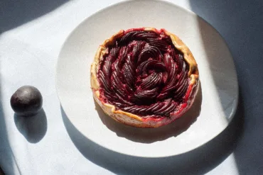 Image of marzipan orange blossom plum galette on light blue tablecloth with scattered fresh plums around it
